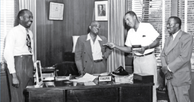 black and white photo of Dr. A.G. Gaston and Dr. E.W. Taggart standing around a desk with two other men
