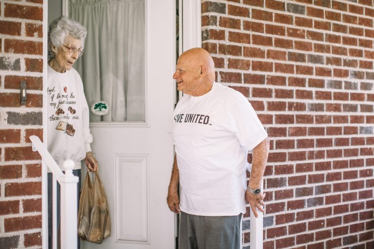 A senior woman stands in her doorway holding a grocery bag, smiling at a United Way Meals on Wheels volunteer wearing a “LIVE UNITED” T-shirt. The two are engaged in friendly conversation on the front porch of a brick house.