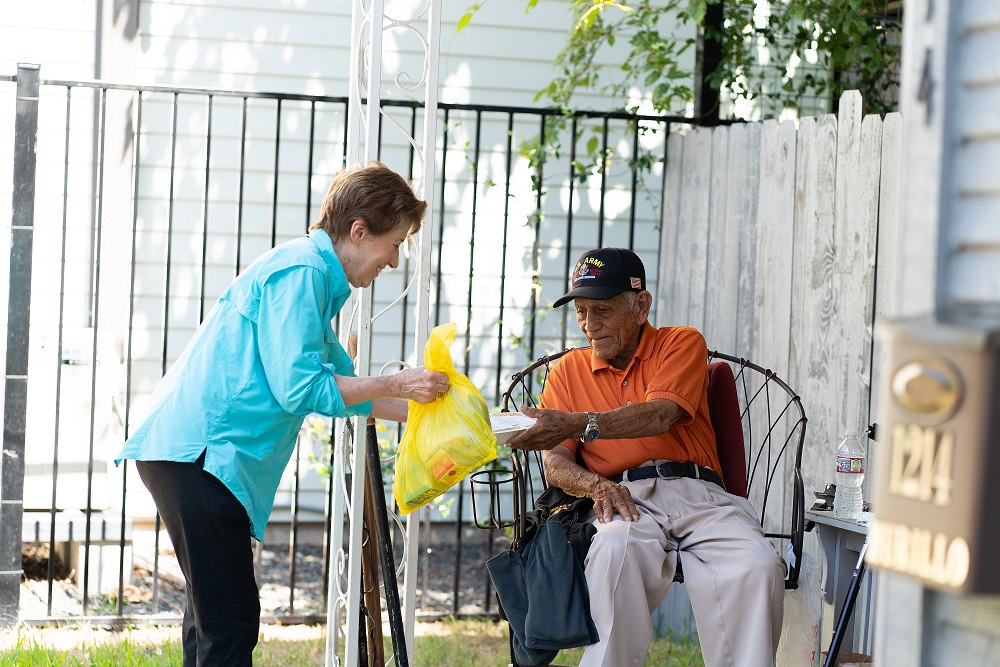 Meals on Wheels volunteer delivers a meal to an older adult seated on his front porch.