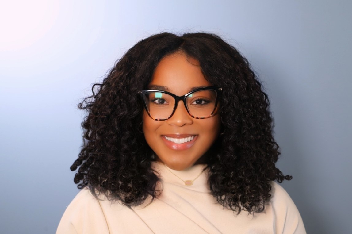 Professional headshot of Aleisha Dunning, a smiling woman with shoulder-length curly hair and dark-rimmed glasses, wearing a light top and small pendant, on a soft blue background; Tutoring Partnership Coordinator in Community Impact.