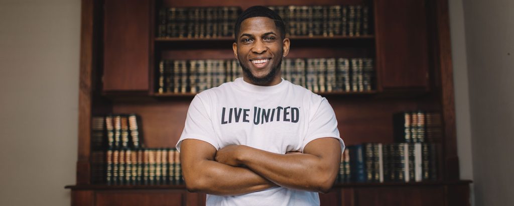 Smiling man wearing a LIVE UNITED T-shirt stands with his arms crossed in front of bookshelves.