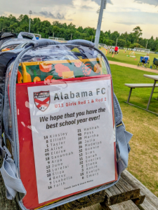 A clear backpack filled with school supplies and a personalized note from Alabama FC sits beside a soccer field.