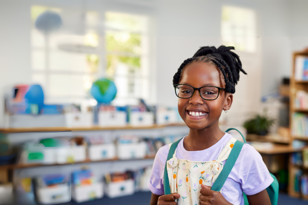 Smiling elementary school student wearing glasses and a backpack, standing in a brightly lit classroom with books and educational materials in the background.