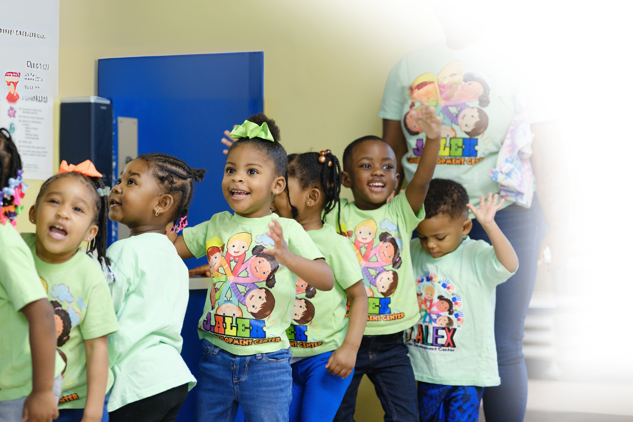 Smiling preschool children dance and raise their hands together in a brightly lit classroom.