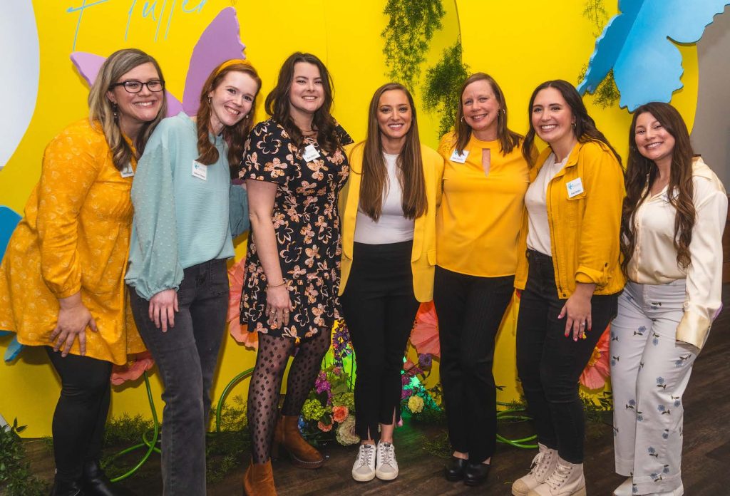 Group of seven women smiling and posing together in front of a colorful yellow backdrop with butterflies and greenery.