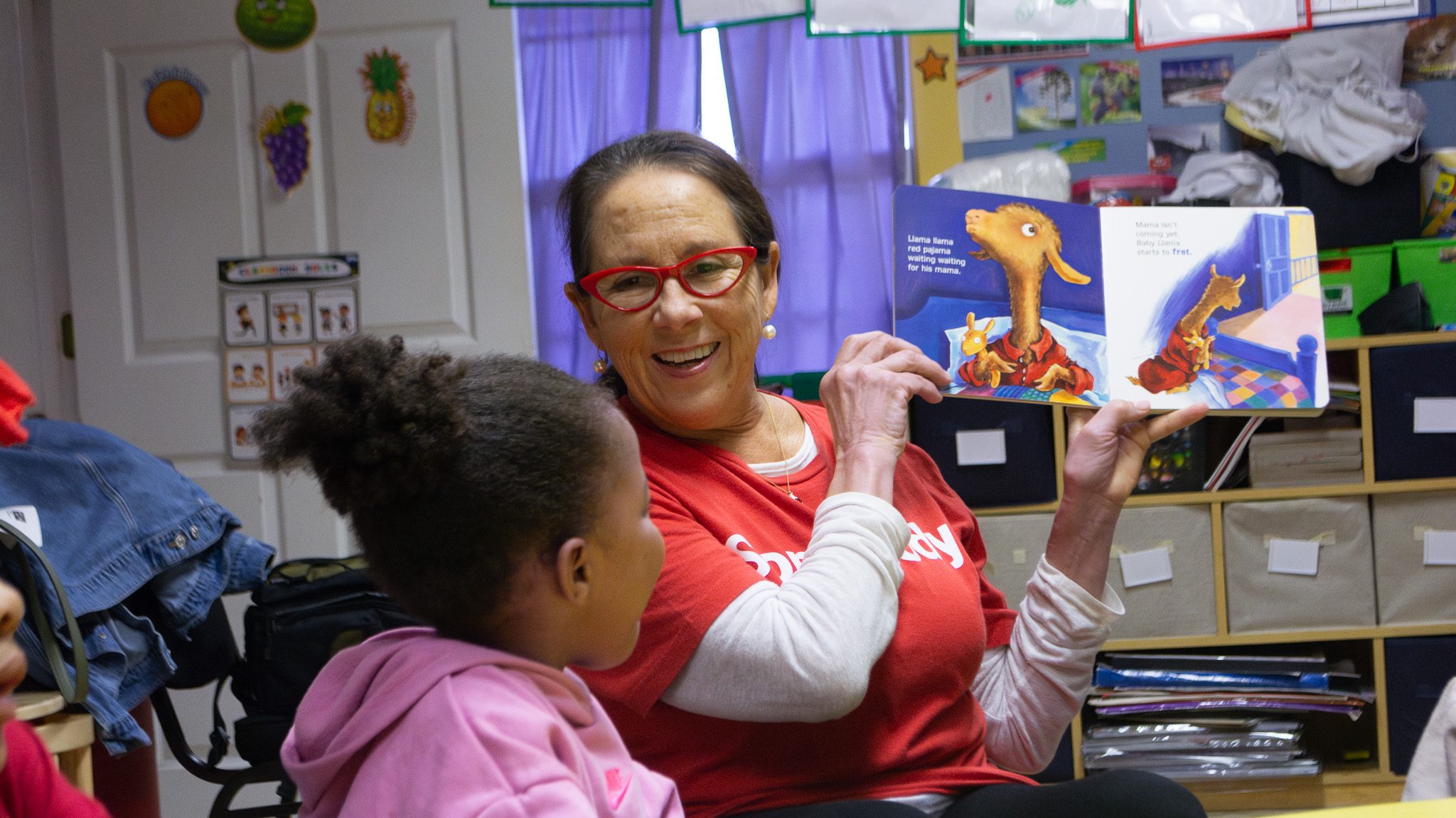 Ladye Franklin, Lead Reading Readiness Specialist with Success by 6, reads a children's book aloud to a young girl in a classroom setting. Wearing red-framed glasses, she smiles warmly while holding up a colorful picture book featuring a llama character.