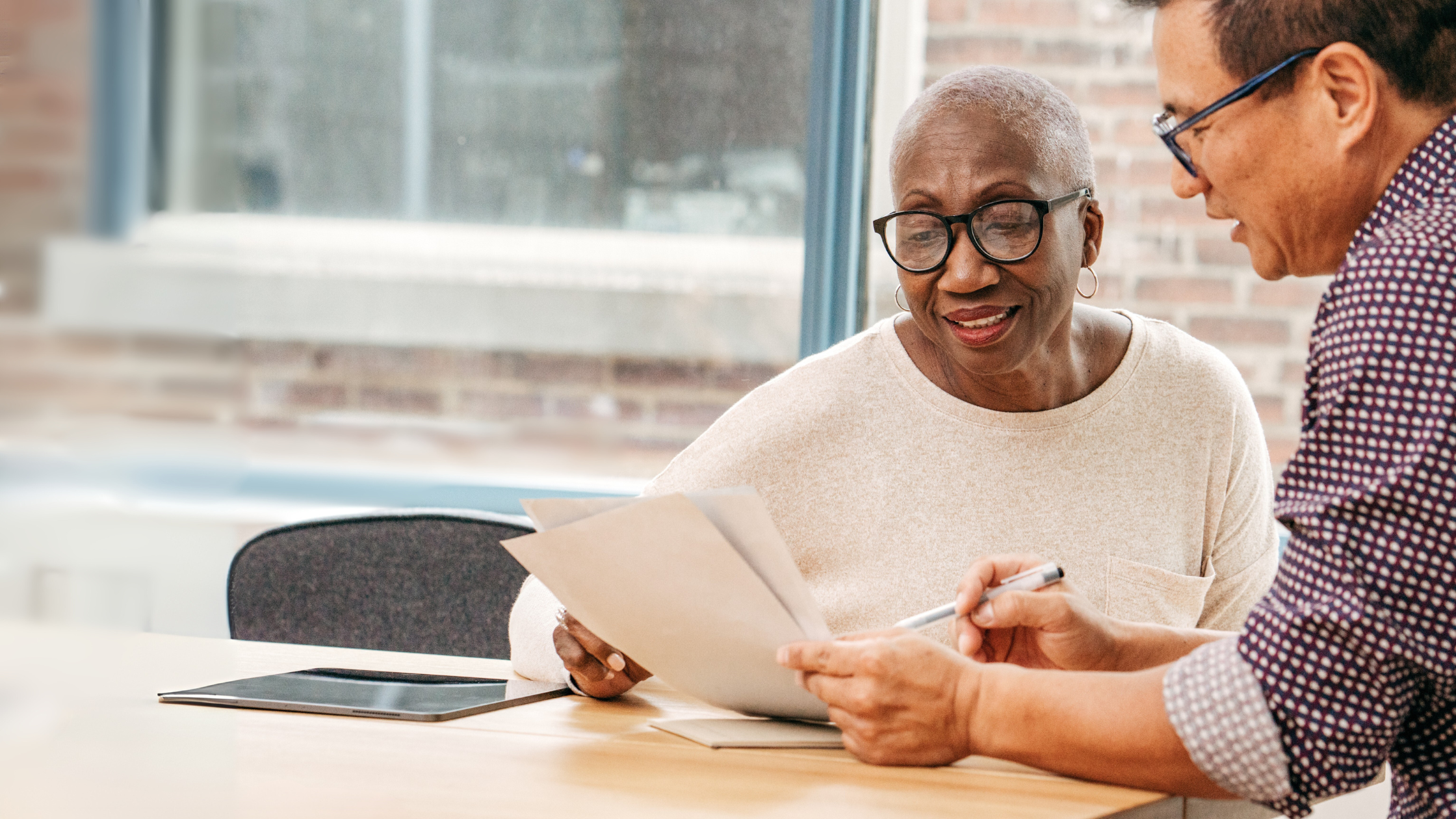 An older woman and a man review financial documents together at a table in a bright office setting. The woman is smiling and holding papers while the man gestures with a pen. A tablet lies on the table nearby