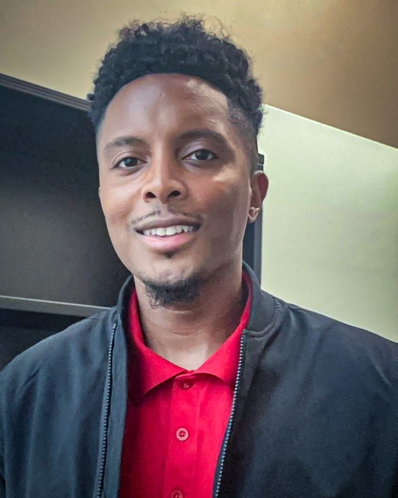 Evan Reddick, a doctoral student at UAB and tutor in United Way’s High-Dosage Tutoring Program, smiles while standing indoors in a red polo and black jacket. He supports sixth-grade students at Huffman Middle School through a partnership between UWCA and Birmingham City Schools.