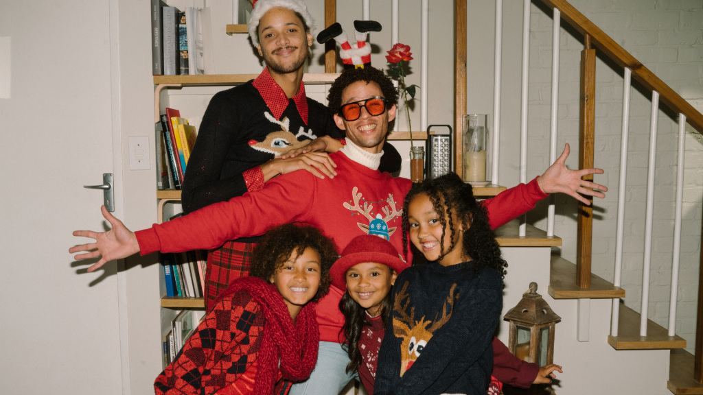 A family in colorful holiday sweaters poses playfully in front of a staircase, smiling and gathered close together.