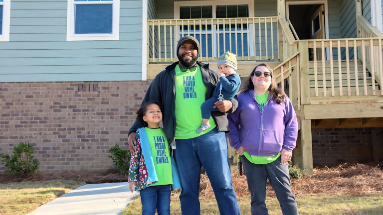 A smiling family of four stands outside a home wearing shirts reading, “I am a proud homeowner.”
