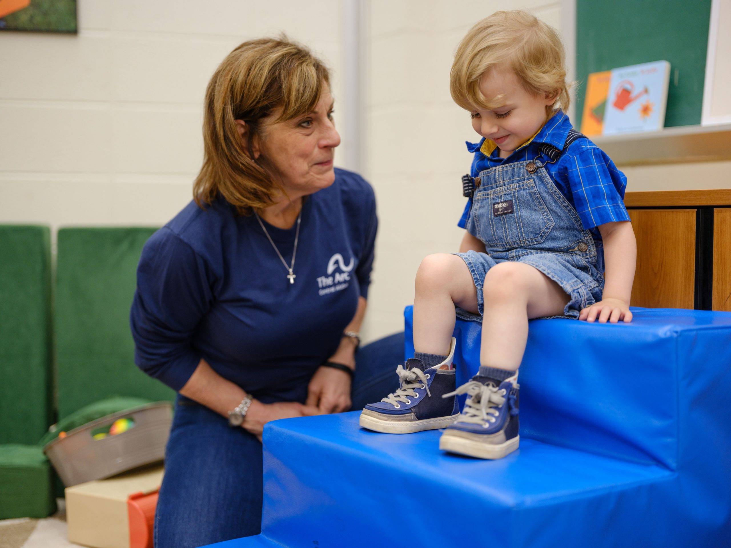 A young boy named Jacob, wearing denim overalls and a blue plaid shirt, smiles as he sits on a soft blue staircase during a therapy session at The Arc of Central Alabama. A staff member kneels beside him, offering encouragement.