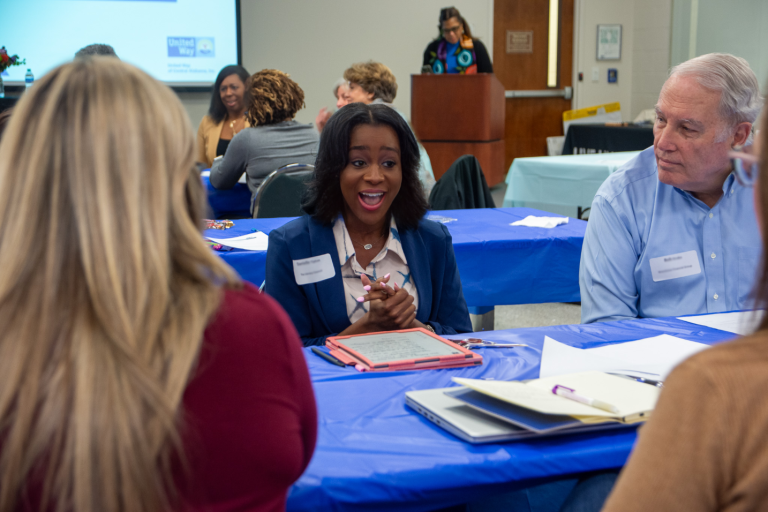 A diverse group of professionals sits around a table covered with a blue tablecloth, engaged in a discussion during a United Way training event. A smiling woman in the center, wearing a name tag, speaks enthusiastically while holding a tablet. Other participants, including an older man and a woman with long blonde hair, listen attentively. In the background, a presenter stands at a podium addressing the room.