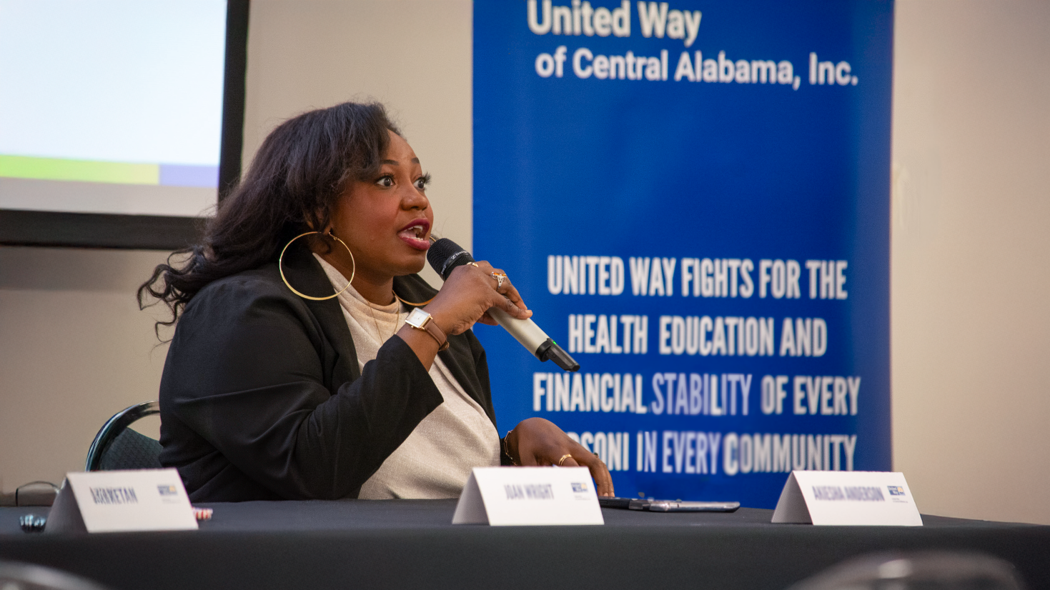 A woman speaks into a microphone while sitting at a panel table during a United Way of Central Alabama event. She wears a black blazer and gold hoop earrings, gesturing as she addresses the audience. A blue United Way banner in the background highlights the organization's mission to support the health, education and financial stability of every person in every community.