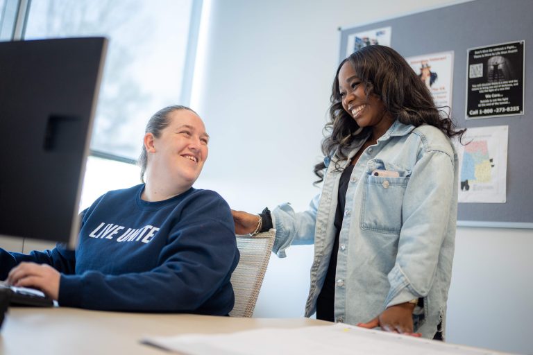 Two smiling women talk beside a computer workstation in a Priority Veteran office.