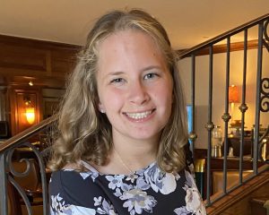Mary White, a young woman with shoulder-length blonde hair, smiles warmly at the camera. She is wearing a black dress with a white and lavender floral print, standing indoors in front of a decorative iron staircase. The setting is warmly lit with wood-paneled walls and ambient lighting.