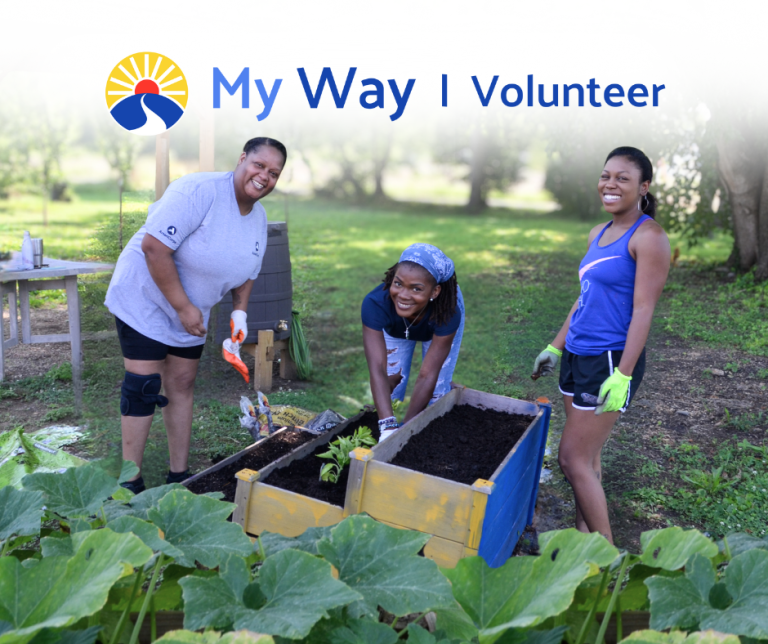 Three My Way volunteers smile while planting vegetables in raised garden beds.