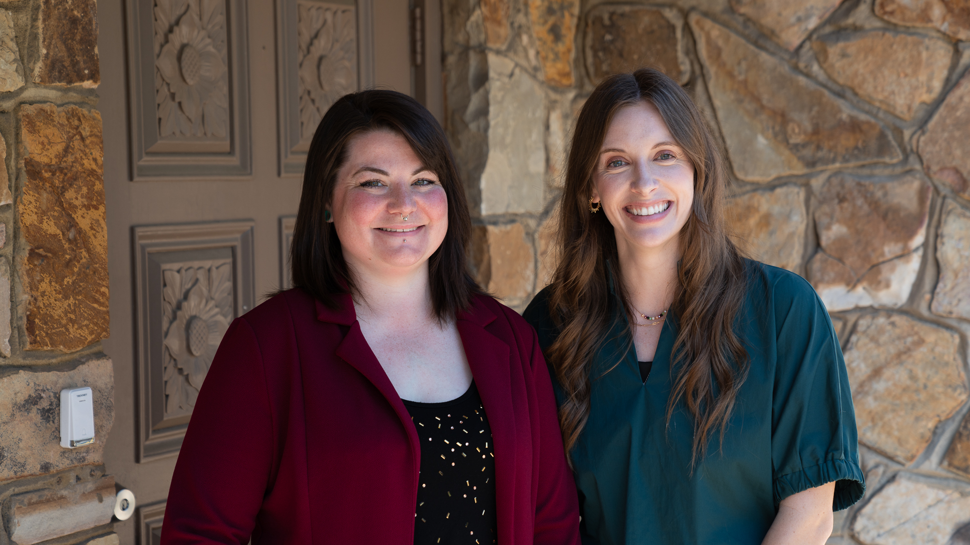 Two women smile while standing side by side outside a stone building, one wearing a burgundy blazer and the other a teal blouse.