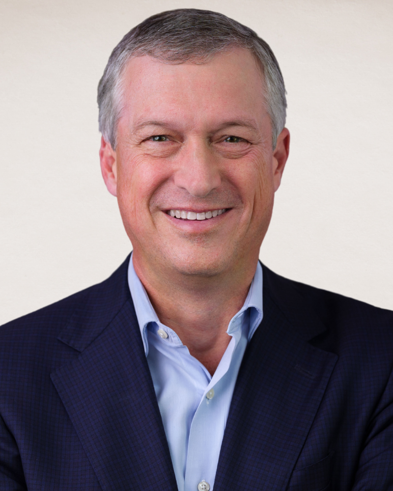 Head-and-shoulders portrait of Scott Adams smiling against a light background, wearing a navy blazer and light blue collared shirt.