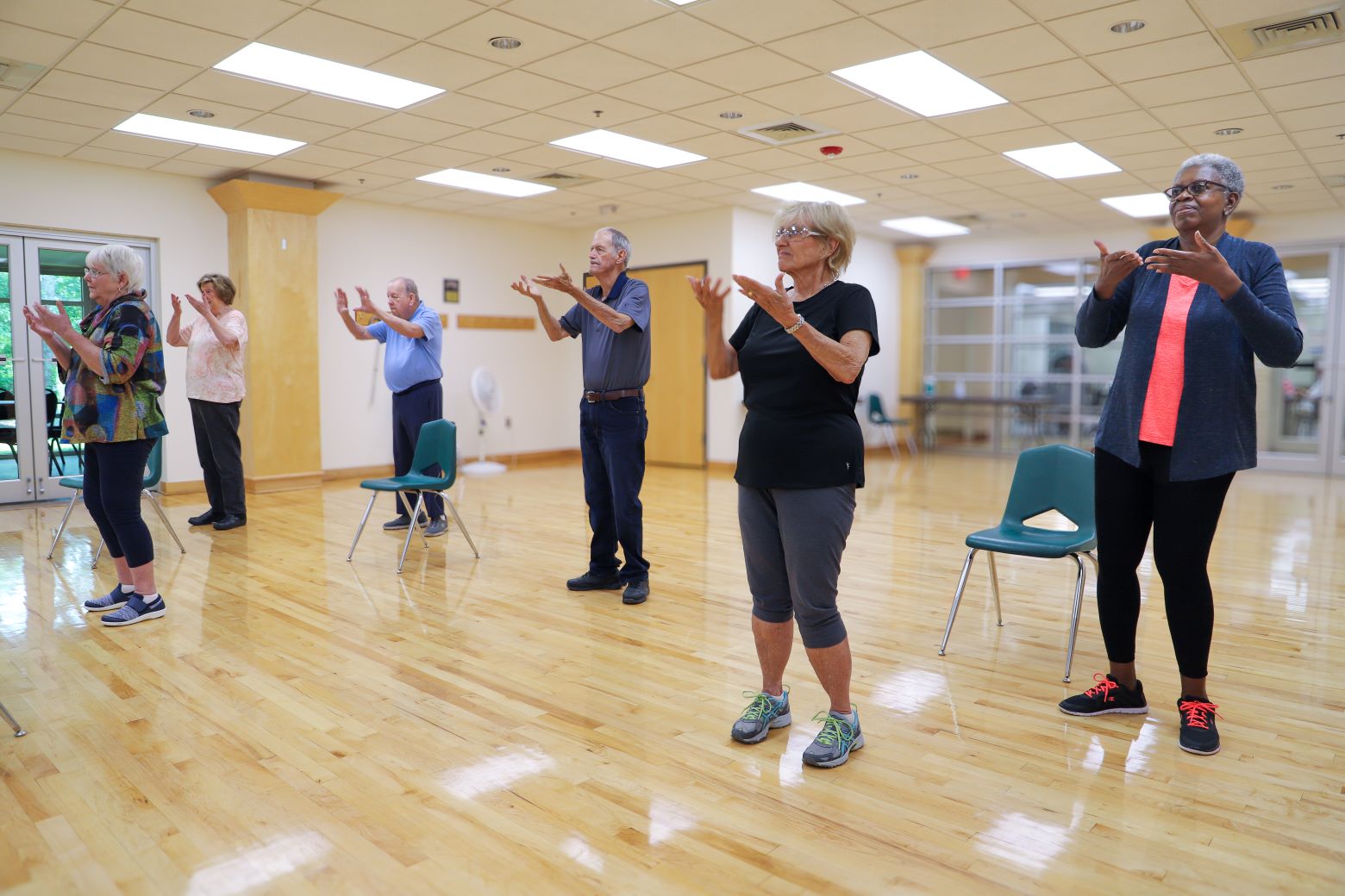 six older adults wearing exercise clothing practice tai chi in a large open room with a wooden floor