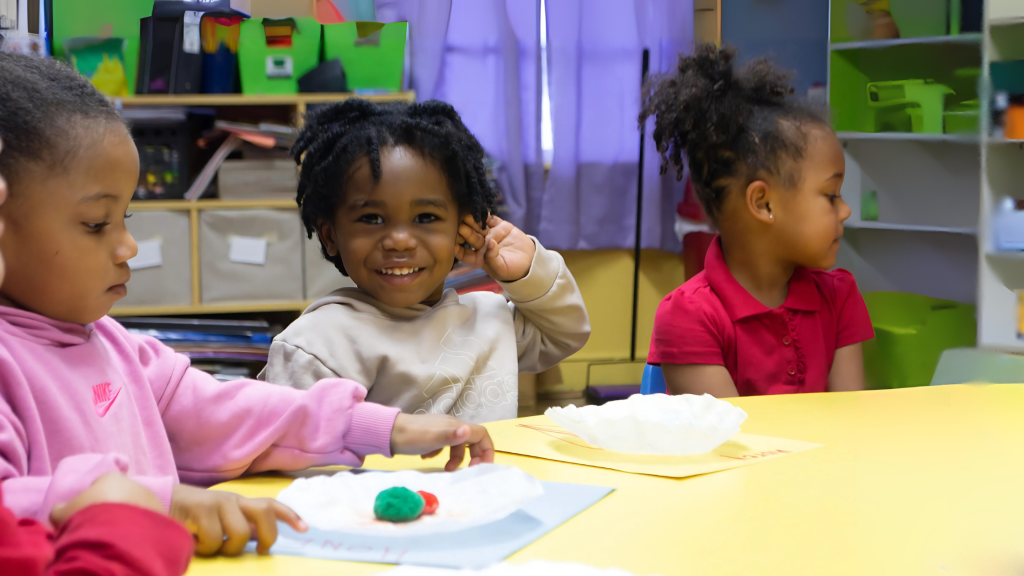 Three young children at a classroom table doing a hands-on art activity with play dough. One child smiles brightly at the camera while others focus on their work.