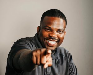 Timothy Alexander, a smiling Black man with close-cropped hair and a trimmed beard, points directly at the camera with a confident and engaging expression. He wears a dark collared shirt and is posed against a simple gray background, radiating warmth and charisma.