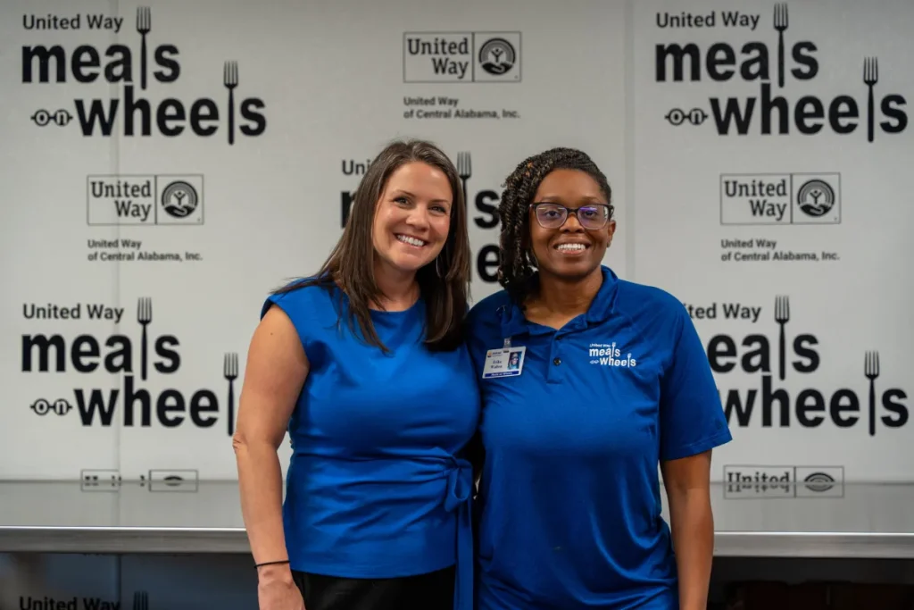 Meals on Wheels Program Director April McMaster and Kitchen Manager Erika Walton smile together in front of United Way Meals on Wheels logos.