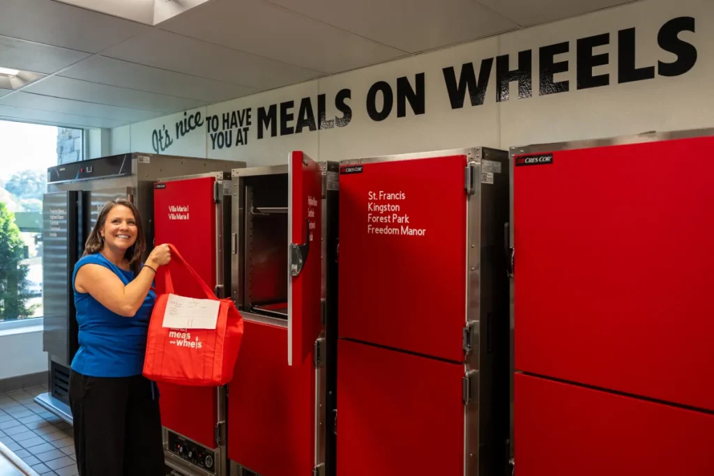 April McMaster holds a red Meals on Wheels delivery bag beside meal-warming cabinets labeled with Jefferson County delivery routes.