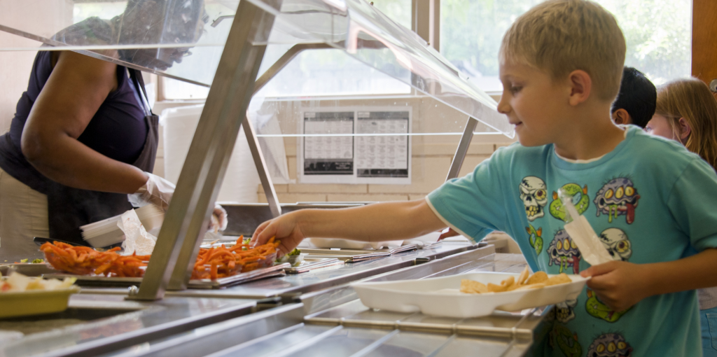 Child receiving a meal tray in a cafeteria line with fresh vegetables being served.