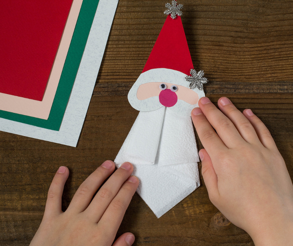 DIY Santa Claus holiday craft made from folded paper with red hat and white beard, created on a wooden table with colored paper sheets.