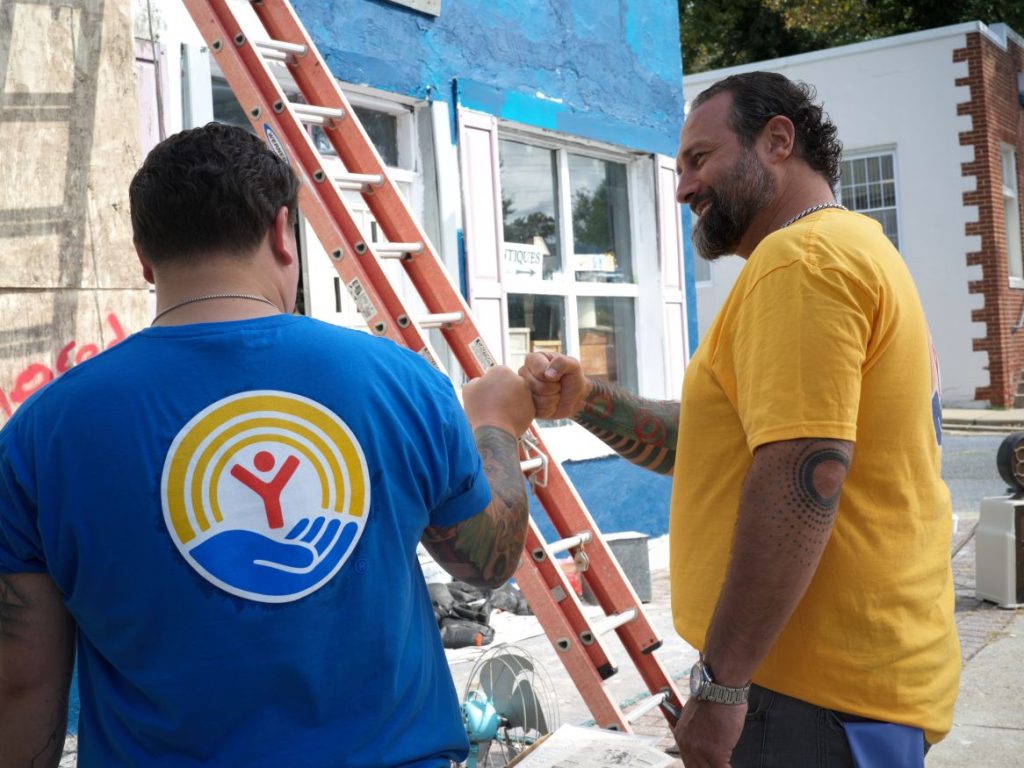 Two volunteers fist-bump beside a ladder while working on an outdoor building improvement project.