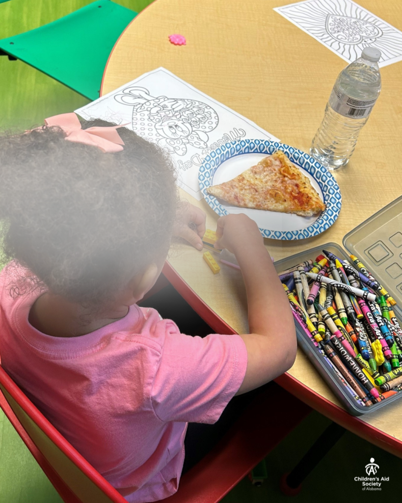 Photo of a young girl wearing a pink t-shirt and matching hairbow seated at a table with crayons, a coloring sheet and a slice of pizza.