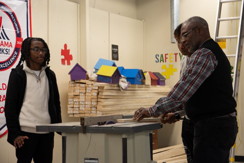 Students and a teacher gather around a workshop table for a demonstration on power tool use.
