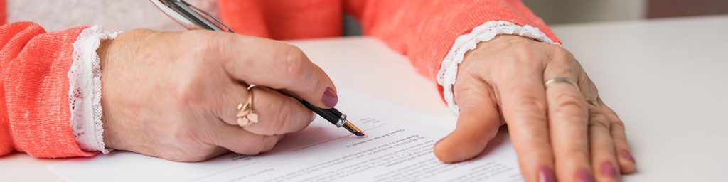 hands of an older person wearing nailpolish and a coral-colored sweater hold a pen and sign a legal document
