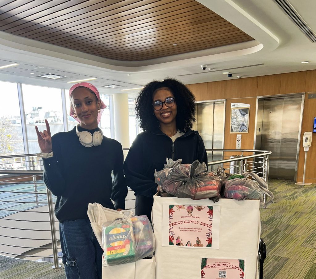 Two UAB students stand beside bins filled with donated period supplies.