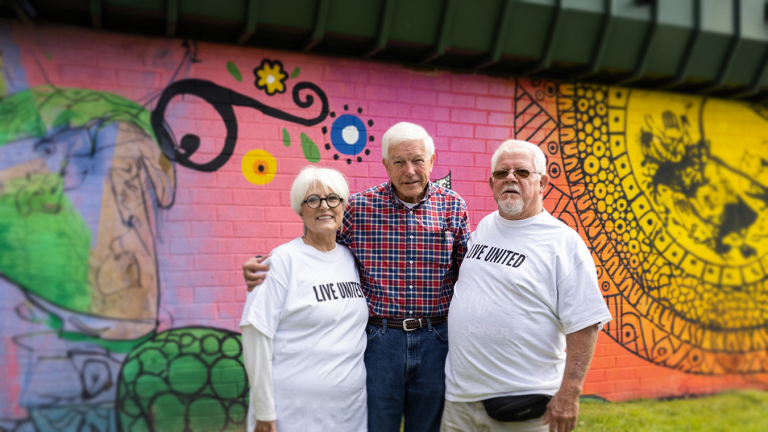 Three older adults stand together in front of a colorful community mural, with two wearing LIVE UNITED shirts.