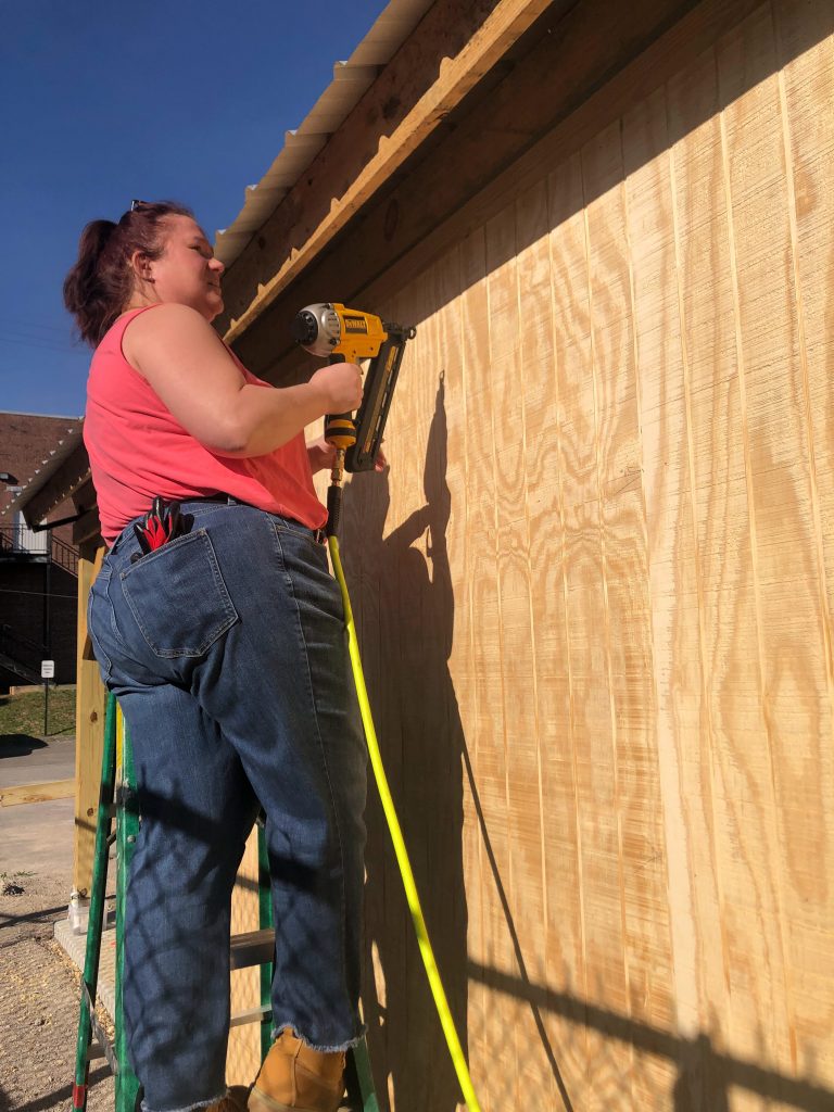 a woman in a ponytail wearing a pink shirt and blue jeans stands on a green ladder holding a yellow power drill