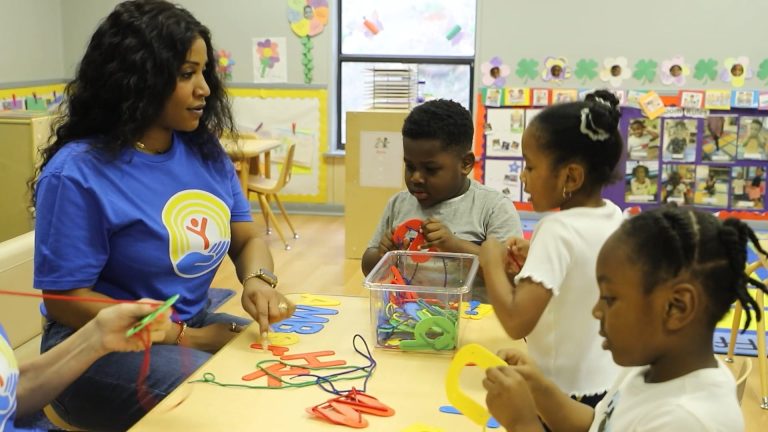 Early learning educator helps three children with a hands-on letter activity in a preschool classroom.